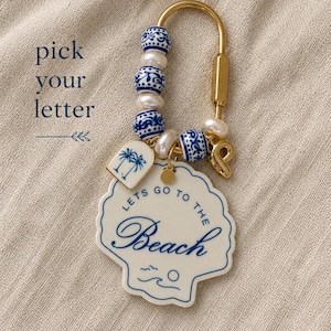 May include: A gold-toned metal keychain featuring blue and white patterned beads, white pearls, and a gold letter P charm. A white shell-shaped pendant displays the text LETS GO TO THE BEACH and a small palm tree charm hangs alongside it.