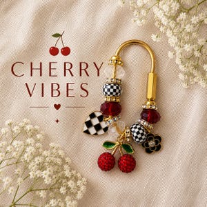 May include: A gold-toned U-shaped keychain featuring a variety of beads including black and white checkered spheres, clear faceted crystals, and deep red beads. A dangling charm shaped like two red cherries with green leaves and a black flower charm.