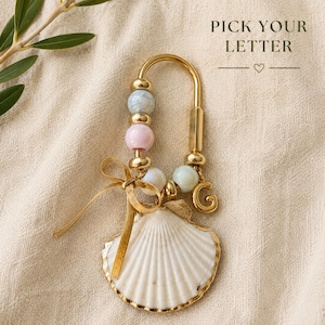 May include: A gold-toned metal keychain featuring a white seashell charm, pastel-colored beads, and a gold letter G charm. A thin gold ribbon is tied to the ring, which is adorned with the text PICK YOUR LETTER with a small heart symbol above.