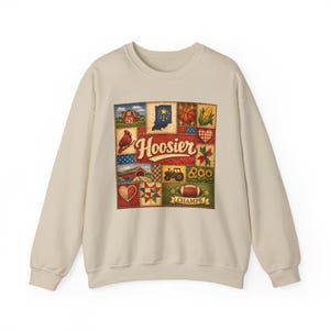 May include: A beige sweatshirt with a patchwork design showcasing Indiana imagery. The design includes a red barn, the state outline, a basketball, corn, a cardinal, a tractor, sunflowers, a football, and the word "Hoosier".