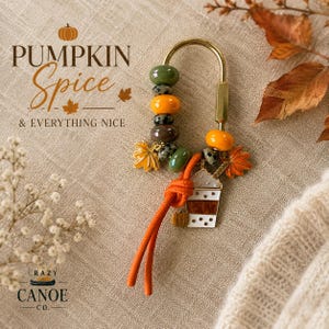 May include: A gold-toned U-shaped keychain featuring a collection of beads in olive green, orange, and speckled brown. The keychain includes metal charms shaped like maple leaves and a coffee cup with white and brown details, plus an orange cord.