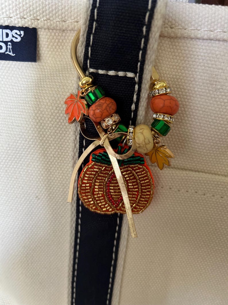 May include: A gold-toned metal keyring adorned with a beaded pumpkin charm, orange and green leaf charms, and various decorative beads in orange, green, and cream. A thin gold ribbon is tied in a bow around the top of the pumpkin charm.
