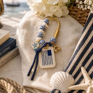 May include: A gold-toned metal keychain featuring blue and white patterned ceramic beads, white pearls, a square charm with a blue starfish illustration, and a rectangular white tag with a blue flag graphic, all accented by a dark blue cord.
