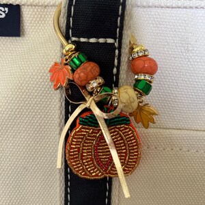 May include: A gold-toned metal bag charm featuring a beaded pumpkin pendant, a thin gold ribbon, and a variety of decorative beads including orange, green, and clear crystal accents, along with small metal maple leaf charms attached to a curved hook.