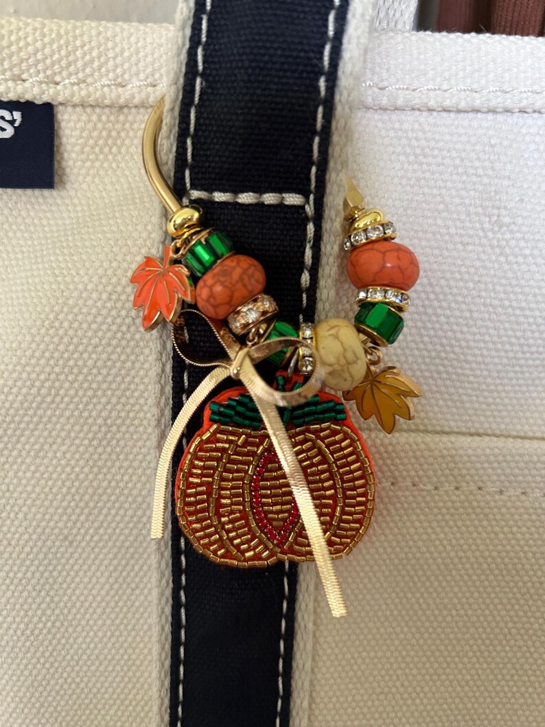 May include: A gold-toned metal bag charm featuring a beaded pumpkin pendant, a thin gold ribbon, and a variety of decorative beads including orange, green, and clear crystal accents, along with small metal maple leaf charms attached to a curved hook.
