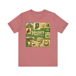 May include: Dusty rose t-shirt featuring a colorful graphic with the words "Hoosier Lucky". The graphic includes images of a cardinal, a barn, a beer mug, a horseshoe, and shamrocks.