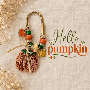 May include: A gold-toned metal keychain featuring a beaded pumpkin charm, orange and green beads, and metal leaf accents. A gold ribbon is tied to the ring. The text "Hello pumpkin" is printed in dark green and orange script next to the keychain.