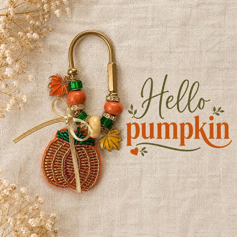 May include: A gold-toned metal keychain featuring a beaded pumpkin charm, orange and green beads, and metal leaf accents. A gold ribbon is tied to the ring. The text "Hello pumpkin" is printed in dark green and orange script next to the keychain.