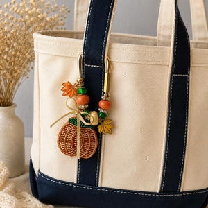 May include: A cream-colored canvas tote bag with navy blue trim and handles. A decorative gold-tone metal clip is attached to the handle, featuring orange and green beads, a gold ribbon bow, and charms shaped like a pumpkin and maple leaves.