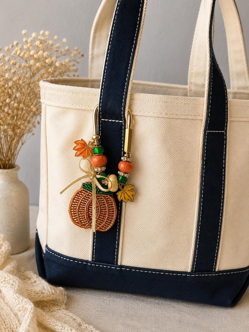 May include: A cream-colored canvas tote bag with navy blue trim and handles. A decorative gold-tone metal clip is attached to the handle, featuring orange and green beads, a gold ribbon bow, and charms shaped like a pumpkin and maple leaves.