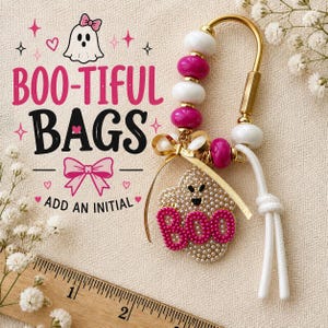 May include: A gold-toned metal keychain featuring alternating white and magenta beads, a gold ribbon bow, and a ghost-shaped charm covered in small white pearls with the word BOO in magenta beads. A wooden ruler shows a scale in inches.