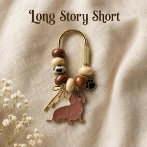 May include: A gold-toned metal keychain featuring the text Long Story Short, adorned with brown, cream, and dalmatian-spotted beads, a small gold bow charm, a black paw print charm, and a brown enamel dachshund dog pendant.
