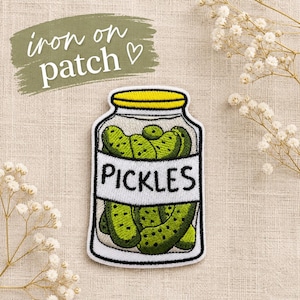 May include: An embroidered iron-on patch featuring a glass jar filled with green pickles. The jar has a yellow lid and a white label with the black text 'PICKLES' printed on it. The patch has a black border and is shaped like a jar.