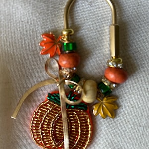 May include: A gold-toned metal keychain featuring a beaded pumpkin charm with green accents, a thin gold ribbon, and various decorative beads in orange, green, and white. The keychain includes two small metal leaf charms in orange and yellow.