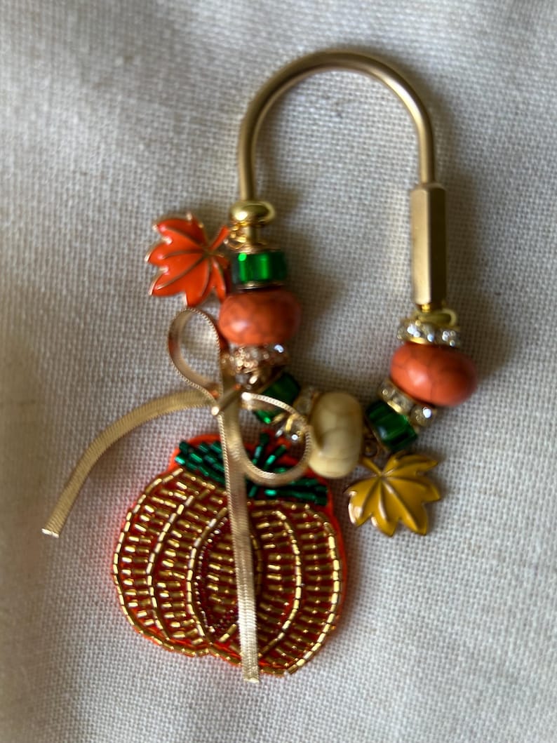 May include: A gold-toned metal keychain featuring a beaded pumpkin charm with green accents, a thin gold ribbon, and various decorative beads in orange, green, and white. The keychain includes two small metal leaf charms in orange and yellow.