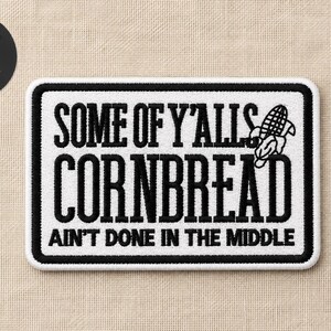 May include: A rectangular iron-on patch with a white background and a thick black border. The patch features black embroidered text that reads SOME OF Y'ALLS CORNBREAD AIN'T DONE IN THE MIDDLE, accompanied by a small black embroidered corn cob icon.
