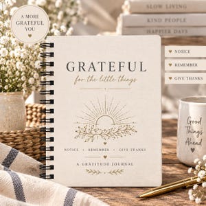 May include: A spiral-bound gratitude journal with a cream-coloured cover featuring the text "GRATEFUL for the little things" and "A GRATITUDE JOURNAL" alongside a gold-toned illustration of a sun rising over leafy branches and small hearts.