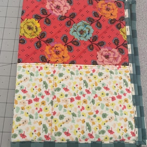 May include: Quilted fabric notebook cover with a floral design. The top section features a red background with large flowers in yellow, pink, and blue. The bottom section has a white background with small colorful flowers. The edges are trimmed with a green and white checkered pattern.