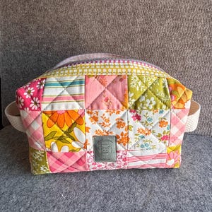 May include: A quilted cosmetic bag with a patchwork design. The bag features a variety of floral and geometric patterns in pink, orange, green, and white. It has a zipper closure and two small fabric handles.
