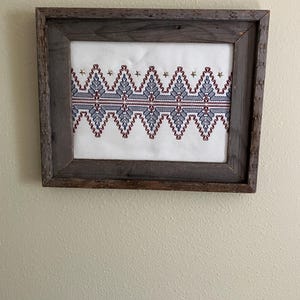 May include: A rectangular cross-stitch embroidery featuring a repeating geometric zigzag pattern in shades of blue and red on a white fabric background, accented with small gold stars, enclosed in a rustic dark brown weathered wooden frame.