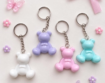 May include: Four bear-shaped keychains in white, lavender, pink, and light blue, each attached to a silver-toned metal split ring and chain. The set is decorated with small pink and purple flower-shaped charms and two translucent butterflies.