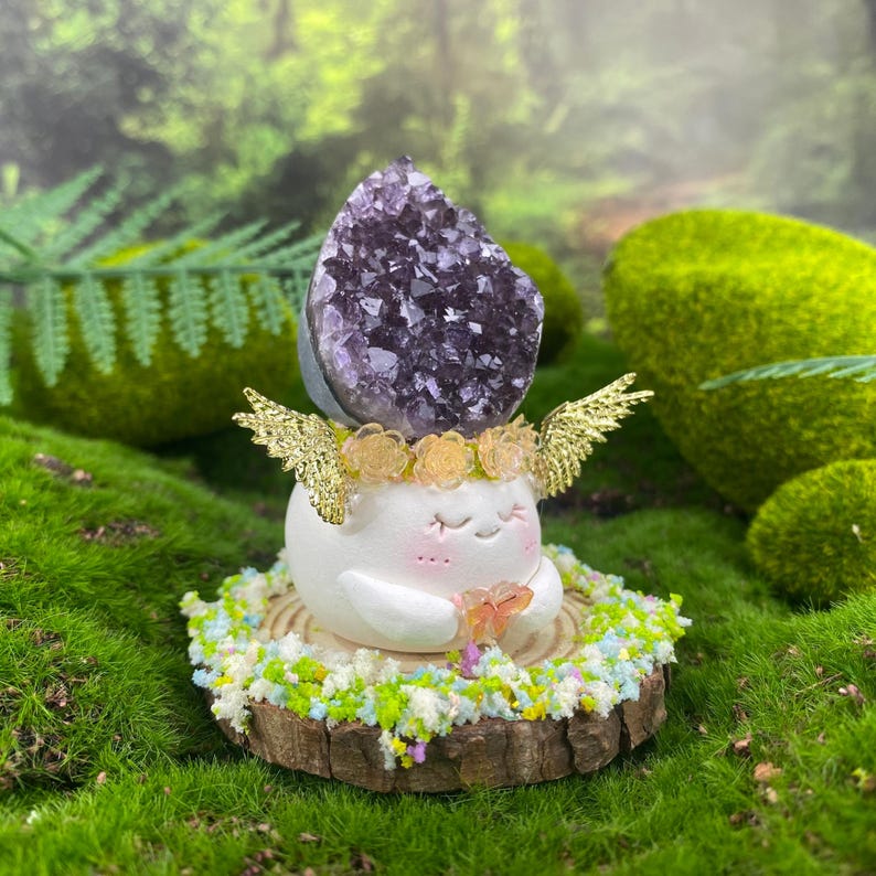 May include: A whimsical figurine featuring a white, smiling character with gold wings and a purple amethyst crystal crown. The figurine rests on a wooden base adorned with colorful floral accents, set against a lush green background.