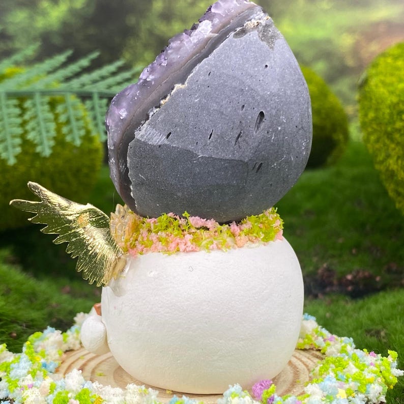 May include: A decorative sculpture featuring a large, dark gray stone with a purple crystal interior atop a white, spherical base. The base is adorned with gold-colored wings and a ring of green and pink decorative elements. The sculpture is set on a wooden slice.