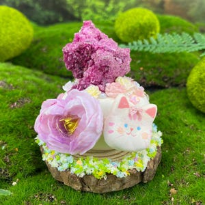 Puede incluir: Una tarta en miniatura artesanal con una flor rosa, una cara de gato y un grupo de cristales rosas. La tarta se asienta sobre una base de madera con detalles florales coloridos, creando una estética de cuento de hadas.