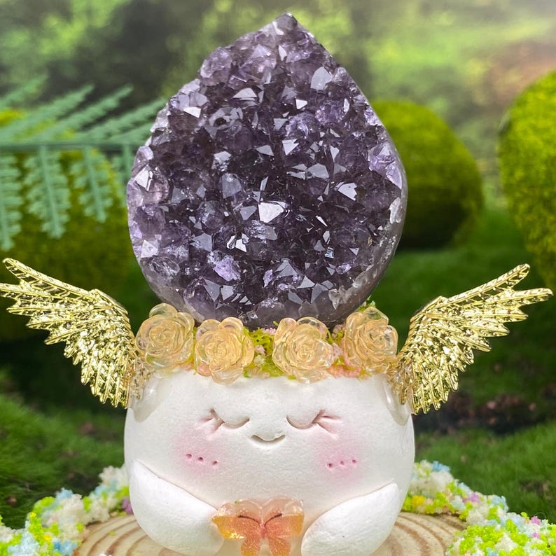 May include: A whimsical figurine featuring a large, dark purple amethyst crystal atop a white, smiling character with gold wings and a floral crown. The figurine holds a small, orange butterfly.
