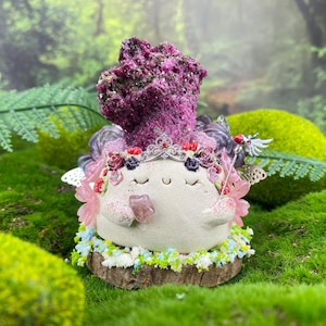 Może przedstawiać: Kapryśna figurka wróżki z kremowym korpusem, ozdobiona koroną, kwiatami i skrzydłami. Wróżka ma duży, teksturowany, magentowy nakrycie głowy. Jest osadzona na drewnianej podstawie otoczonej małymi kwiatami.