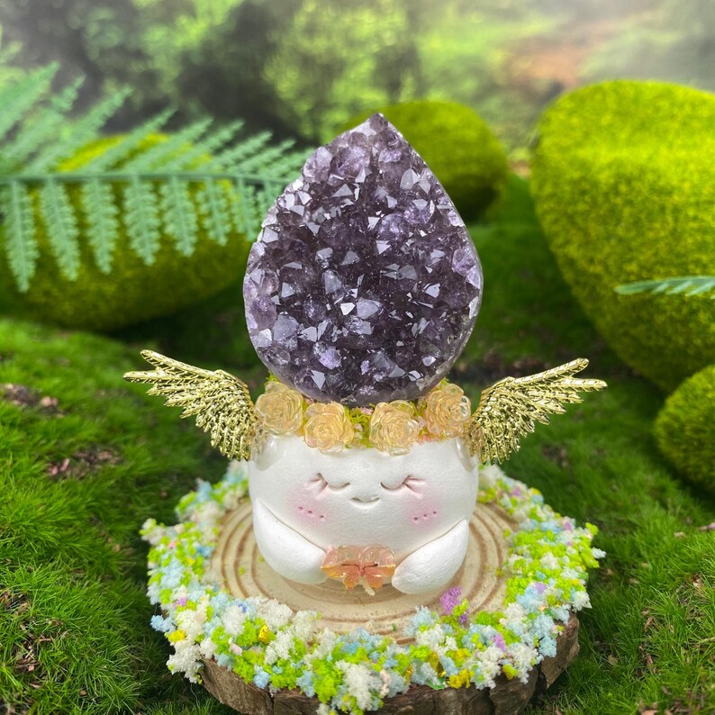 May include: A whimsical figurine featuring a large, dark purple amethyst crystal atop a white, smiling character with golden wings and a floral crown. The figurine rests on a wooden slice surrounded by colorful moss and flowers.