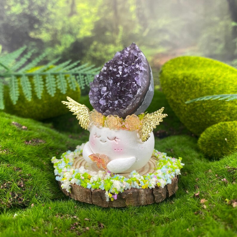 May include: A whimsical figurine featuring a white, smiling character with a purple amethyst crystal headpiece, gold wings, and a floral crown. It rests on a wooden base adorned with colorful floral accents, set against a backdrop of green moss and foliage.