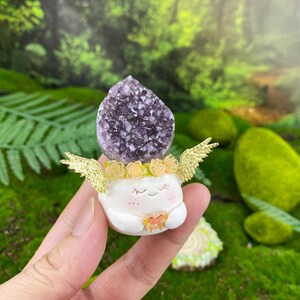 May include: A whimsical figurine featuring a white, smiling character with closed eyes, adorned with gold wings and a floral crown. A large, purple amethyst crystal sits atop the character's head. The figurine has a small, orange butterfly on its chest.