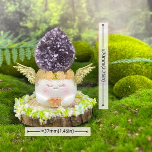 May include: A whimsical figurine featuring a smiling white face with closed eyes, gold wings, and a crown of yellow roses. The figurine is topped with a large purple amethyst crystal and sits on a wooden base with green and white floral accents. The figurine is approximately 2.75 inches tall and 1.46 inches wide.