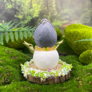 May include: A whimsical fairy figurine with a gray stone head, white body, and gold wings. The fairy is adorned with a floral collar and sits on a wooden base surrounded by colorful floral accents. The background features green moss and foliage.