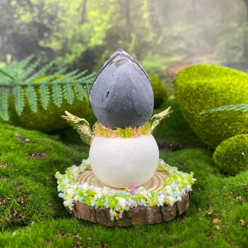 May include: A whimsical fairy figurine with a gray stone head, white body, and gold wings. The fairy is adorned with a floral collar and sits on a wooden base surrounded by colorful floral accents. The background features green moss and foliage.