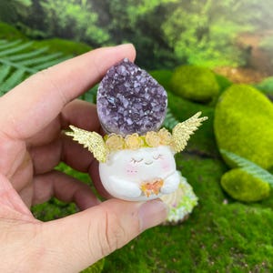May include: A whimsical figurine featuring a white, smiling character with a purple amethyst crystal head. It has gold-colored wings and a floral crown. The character is holding a small, orange butterfly. The figurine is set against a backdrop of green moss.