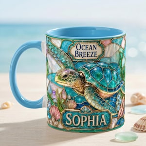 Può includere: Una tazza in ceramica con interno e manico azzurro, caratterizzata da un'illustrazione in stile vetrata raffigurante una tartaruga marina circondata da conchiglie. Il testo Ocean Breeze è stampato in alto e il nome Sophia su un banner in basso.