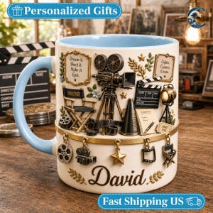 May include: A cream-colored ceramic mug with a light blue interior and handle. The exterior features 3D-style film production motifs including a camera, director chair, and clapperboard. The name David is printed in script, with hanging charms.