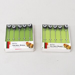 May include: Two packages of black and white 8-ball shaped food picks. Each package contains 5 picks and has a green background with the text "Shenfeld Variety Picks includes 5 picks".
