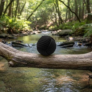 May include: A black decorative ball made of tightly woven rope, resting on a weathered tree branch. The ball is spherical and has a textured surface. The background features a flowing stream and lush green forest.