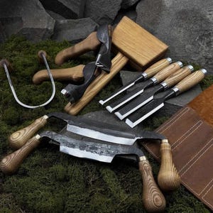 May include: A collection of woodworking tools including two drawknives with wooden handles, three chisels with wooden handles, a wooden mallet, a metal U-shaped tool, and a brown leather tool roll. The tools feature dark metal blades and light wood.