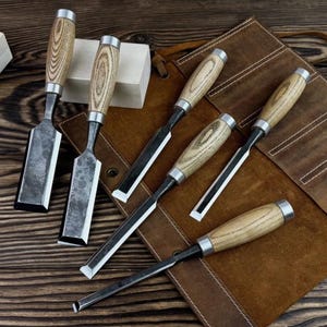 May include: A set of six wood chisels with light-colored wooden handles and silver metal ferrules. The steel blades vary in width and length, featuring a dark, textured finish. The tools are arranged on a brown leather roll-up storage case.