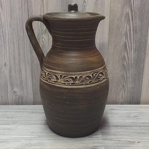 May include: A dark brown ceramic pitcher with a lid and handle. The pitcher has a decorative band of carved floral designs around the middle. The surface has horizontal lines. The pitcher is on a wooden surface.