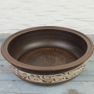 May include: A round, brown ceramic bowl with a decorative, cream-colored floral pattern around the rim. The interior of the bowl is a darker brown, and the rim is slightly raised. The bowl sits on a light blue wooden surface.