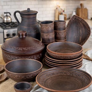 Op de afbeelding: Een verzameling handgemaakt, donkerbruin keramisch servies. De set bevat een grote kan, kommen, borden en een ovenschotel met deksel, allemaal met decoratieve bloempatronen. Het rustieke aardewerk is geplaatst op een neutrale ondergrond.