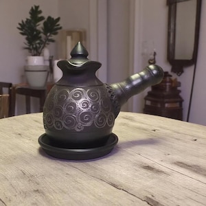 Könnte beinhalten: Eine dunkelgraue türkische Kaffeekanne aus Keramik mit langem Ausguss und kleinem, rundem Boden. Die Kanne hat ein dekoratives Wirbelmuster in einer helleren Graufärbung. Die Kanne steht auf einer runden, dunkelgrauen Untertasse.