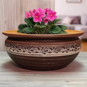 Op de afbeelding: Een bruine keramische bloempot met een decoratieve crèmekleurige band en een brede rand. Het bevat een Afrikaanse viooltje met helder roze bloemen en groene bladeren. De pot staat op een licht houten oppervlak.