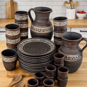 Puede incluir: Un juego de vajilla de cerámica marrón rústico con platos, tazas y jarras. El juego incluye una pila de platos, varias tazas y dos jarras. Cada pieza tiene una banda floral decorativa blanca y negra.