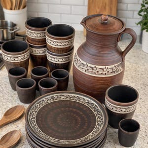 May include: A rustic brown ceramic dinnerware set. The set includes a large jug with a lid, plates, cups, and small shot glasses. Each piece has a decorative white and brown floral pattern around the rim.